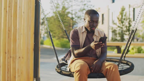 Hombre-Negro-Sentado-Usando-Un-Teléfono-Inteligente-En-El-Columpio-Al-Aire-Libre